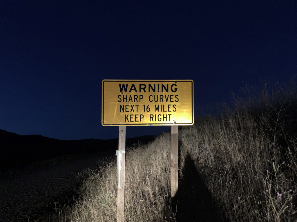 A yellow warning sign for sharp curves and narrow miles photographed after dark.