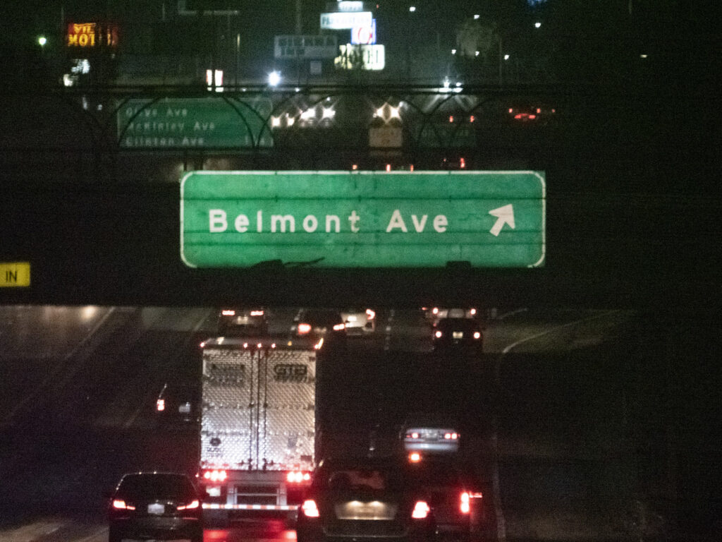 An overhead Belmont Avenue freeway sign photographed in nighttime traffic.