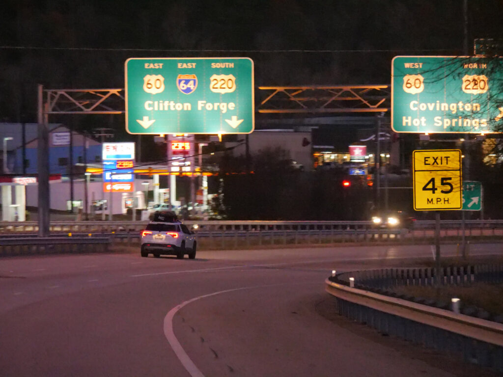Overhead interstate guide signs direct traffic toward Clifton Forge and Saginaw at night.