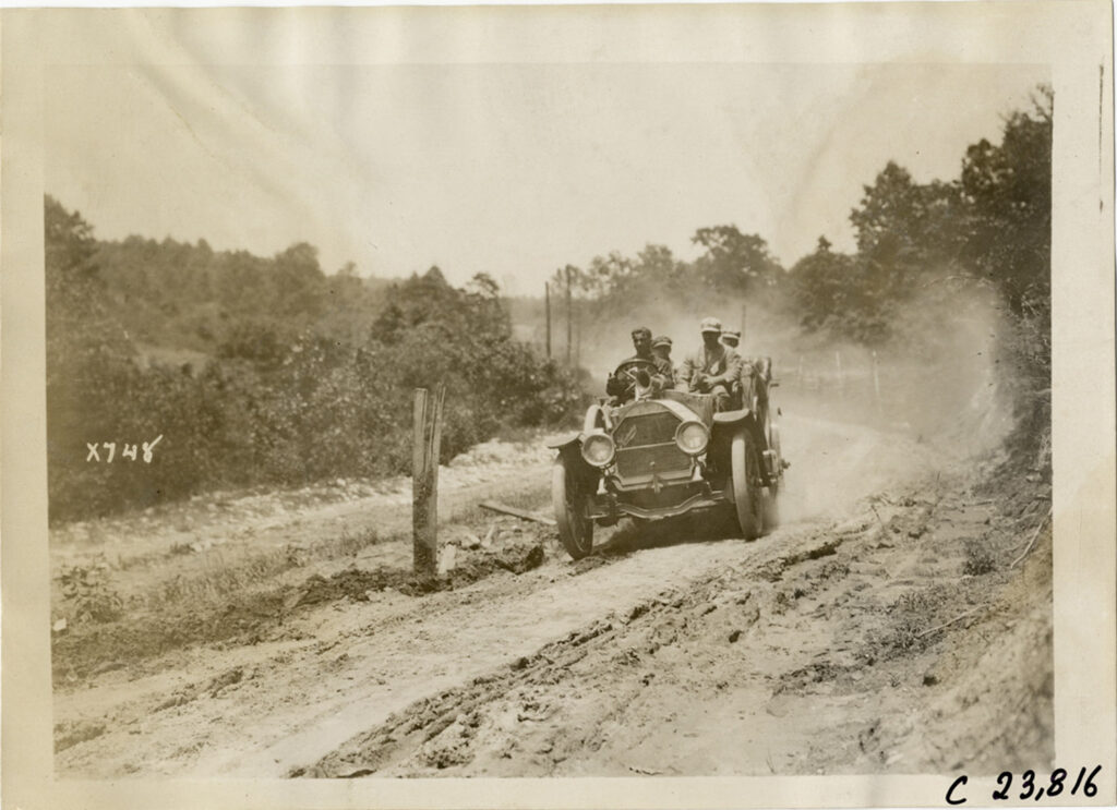 Early automobile driving on an unpaved road with dust behind it.